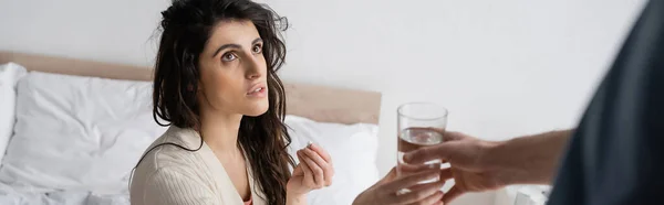 Donna bruna che tiene le pillole e prende un bicchiere d'acqua dal fidanzato offuscato in camera da letto, banner — Foto stock