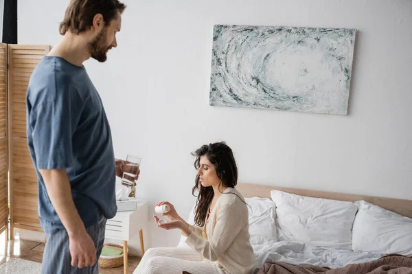 Fürsorglicher Mann hält Glas Wasser neben Freundin mit Tabletten im Schlafzimmer — Stockfoto