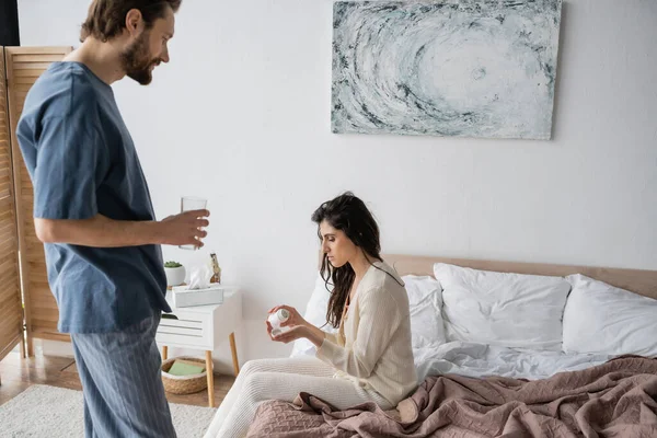 Side view of woman holding pills near caring boyfriend with glass of water in bedroom — Stock Photo