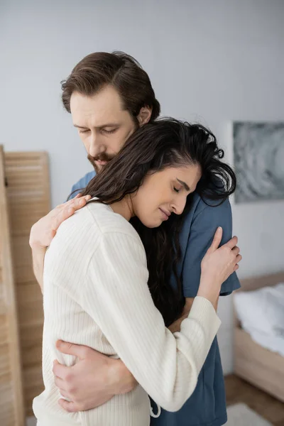Bearded man hugging and calming displeased girlfriend in bedroom at home — Stock Photo