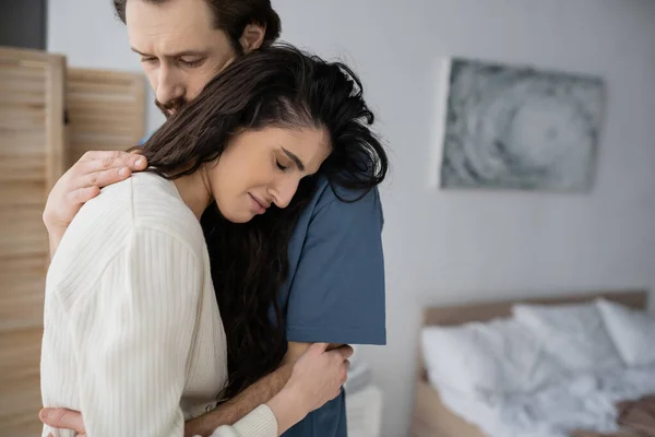 Hombre cariñoso abrazando a novia llorando en el dormitorio borroso en casa - foto de stock