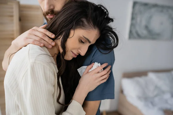Bärtiger Mann beruhigt traurige Freundin mit Serviette im verschwommenen Schlafzimmer — Stockfoto
