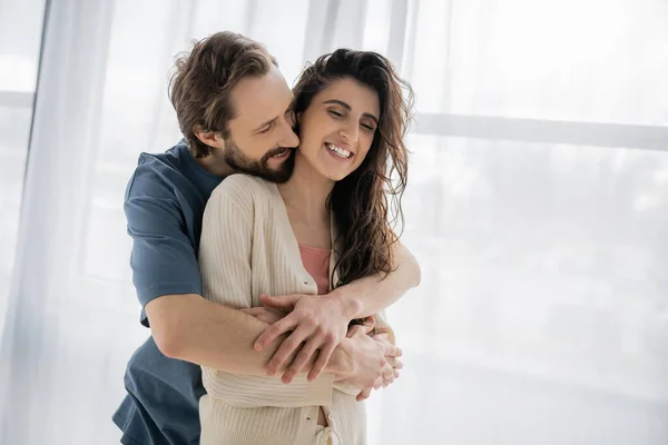 Bearded man embracing cheerful brunette girlfriend at home — Stock Photo