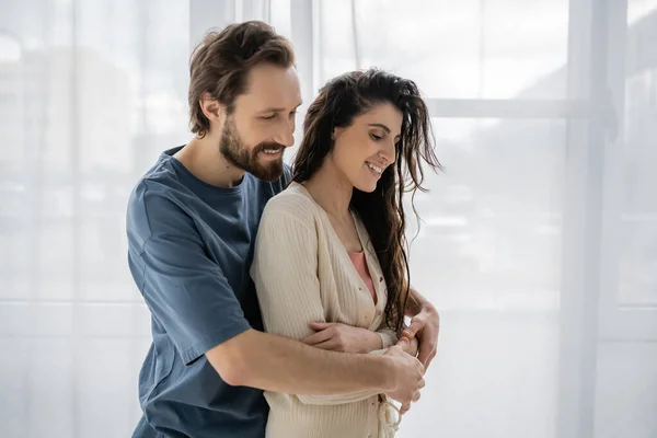 Sonriente hombre abrazando morena novia en ropa casual en casa - foto de stock