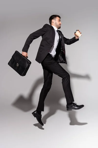 Vista lateral de hombre de negocios con estilo en traje negro levitando con café para llevar y maletín de cuero sobre fondo gris - foto de stock