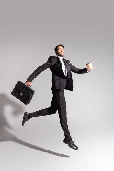 Vista lateral del hombre de negocios encantado con taza desechable y maletín levitando sobre fondo gris - foto de stock