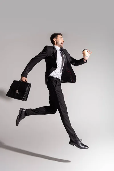 Вид збоку схвильованого бізнесмена в чорному костюмі, вирівнюється зі шкіряним портфелем і паперовою чашкою на сірому фоні — Stock Photo