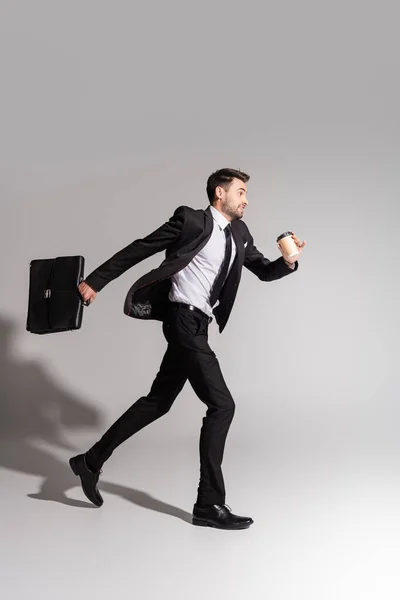 Боковой вид положительного бизнесмена в черном костюме, бегущего с портфелем и бумажной чашкой на сером фоне — стоковое фото
