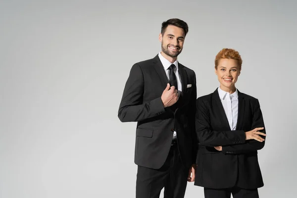 Happy business partners in black stylish suits looking at camera isolated on grey — Stock Photo