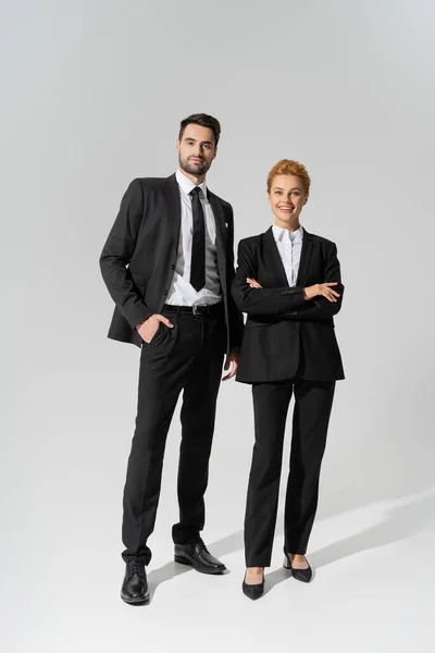 Longitud completa de colegas de negocios sonrientes en negro y elegante ropa formal sobre fondo gris - foto de stock