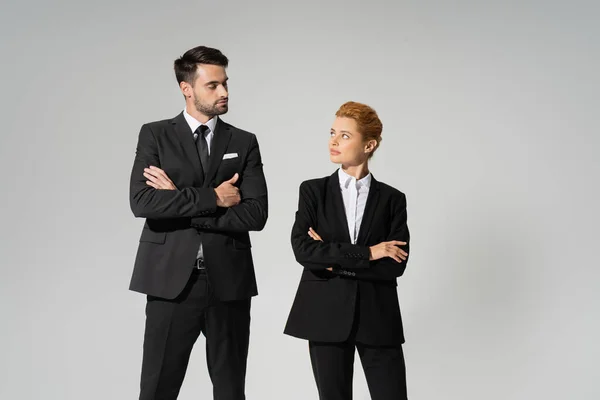 Gente de negocios escépticos en trajes negros de pie con los brazos cruzados y mirándose unos a otros aislados en gris - foto de stock