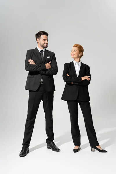 Comprimento total em colegas de negócios alegres em pantsuits elegantes sorrindo para cada um enquanto de pé com os braços cruzados no fundo cinza — Fotografia de Stock