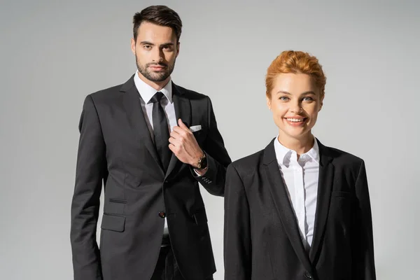Cheerful redhead businesswoman smiling at camera near confident businessman in stylish suit isolated on grey — Stock Photo