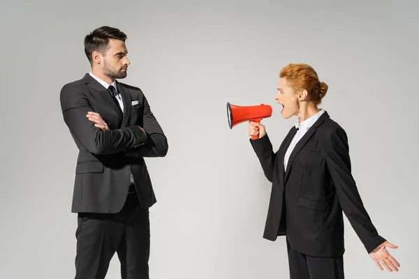 Mujer de negocios enojada gritando en megáfono a hombre confiado en traje de pie con los brazos cruzados aislados en gris - foto de stock