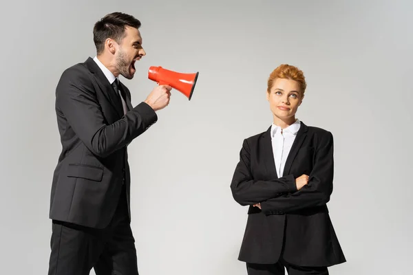 Angry businessman screaming in loudspeaker near skeptical woman standing with crossed arms isolated on grey — Stock Photo