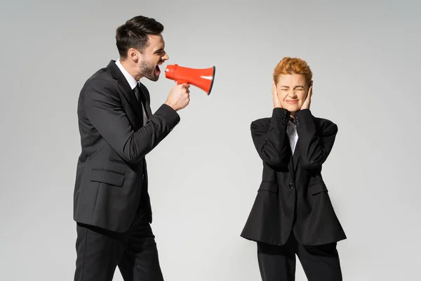 Redhead manager with closed eyes covering ears with hands near boss screaming in megaphone isolated on grey — Stock Photo