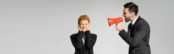 Redhead woman with closed eyes covering ears with hands near irritated boss screaming in megaphone isolated on grey, banner — Stock Photo