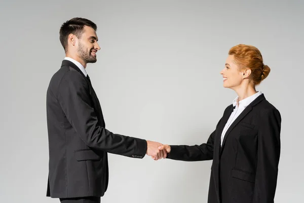 Side view of joyful business partners in black blazers shaking hands isolated on grey — Stock Photo