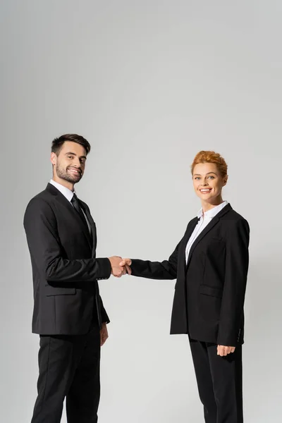 Успешных деловых партнеров в черных пиджаках пожимающих руки и смотрящих в камеру, изолированную на сером — стоковое фото