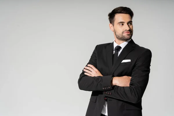 Улыбающийся бизнесмен в черном костюме, стоящий со сложенными руками и смотрящий в сторону изолированный на сером — стоковое фото