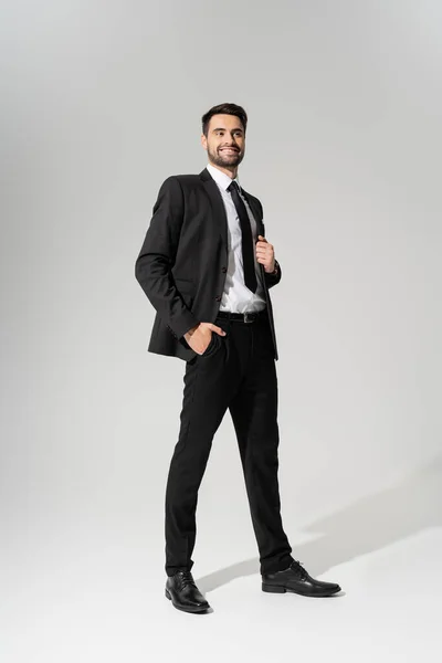 Auf fröhlichem Geschäftsmann in schwarzem Hosenanzug, der mit der Hand in der Tasche posiert und auf grauem Hintergrund wegschaut — Stockfoto