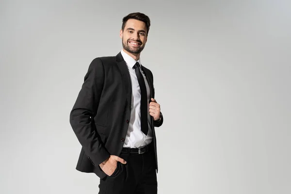 Bärtiger Geschäftsmann im schwarzen Anzug, die Hand in der Tasche und lächelnd in die Kamera isoliert auf grau — Stockfoto
