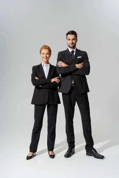Full length on successful business partners in black suits crossing arms and looking at camera on grey background — Stock Photo
