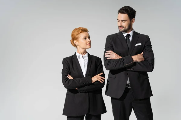 Colleghi di lavoro positivi in blazer neri in piedi con le braccia incrociate e guardarsi isolati su grigio — Foto stock