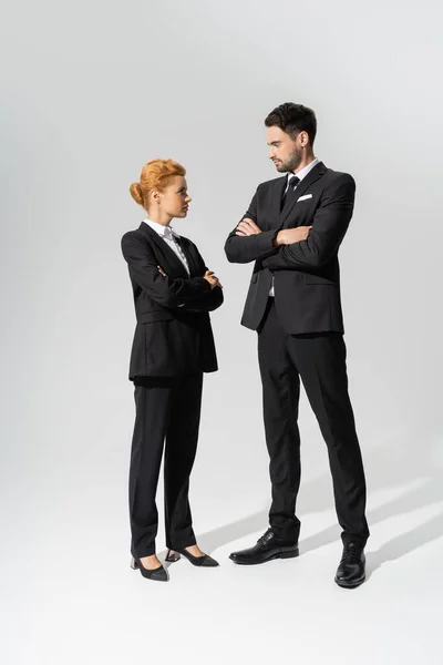 In voller Länge blicken skeptische Geschäftsleute in schwarzen Anzügen einander an, während sie mit verschränkten Armen vor grauem Hintergrund stehen. — Stockfoto