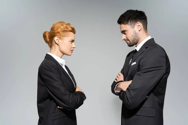 Side view of angry business people looking at each other while standing with crossed arms isolated on grey — Stock Photo