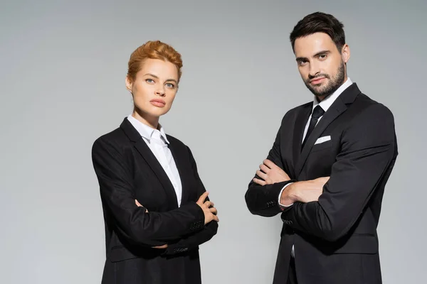 Confident business colleagues in black blazers standing with folded arms and looking at camera isolated on grey — Stock Photo