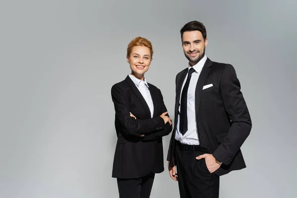 Successful and stylish business partners in black suits smiling at camera isolated on grey — Stock Photo