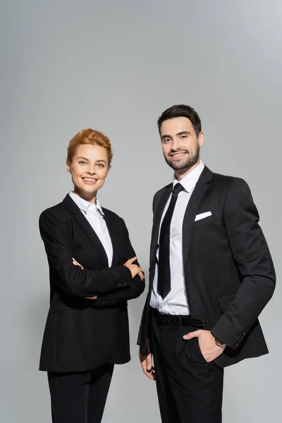 Zufriedene Geschäftskollegen in schwarzen schicken Anzügen, die in die Kamera blicken und vereinzelt auf grau lächeln — Stockfoto