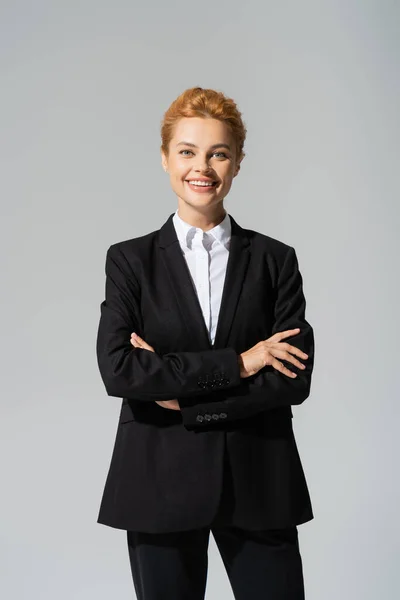 Alegre mujer de negocios en blazer negro sonriendo a la cámara mientras posa con los brazos cruzados aislados en gris - foto de stock