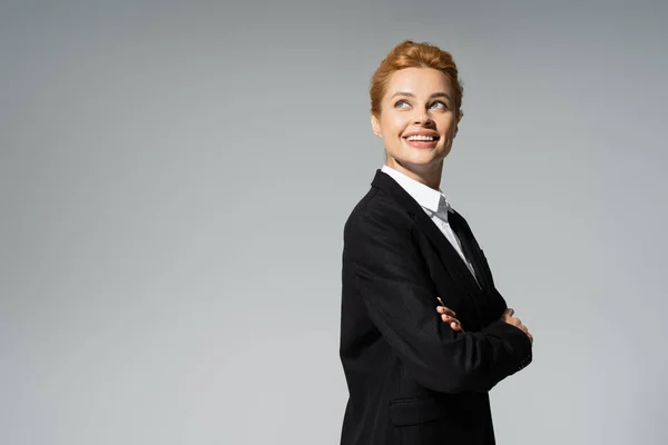 Happy redhead businesswoman in black blazer looking away while standing with crossed arms isolated on grey — Stock Photo