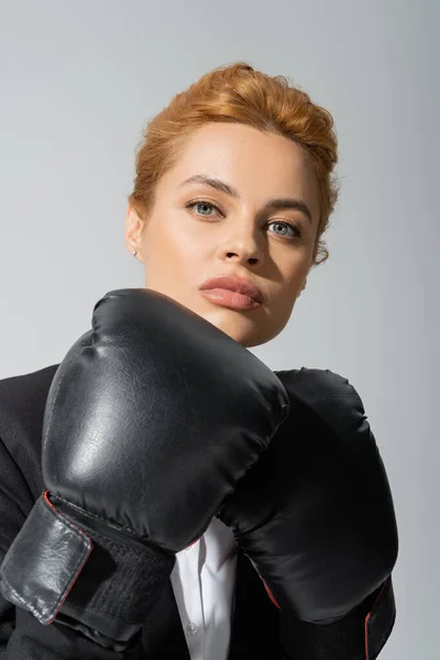 Retrato de mujer de negocios pelirroja confiada en guantes de boxeo mirando a la cámara aislada en gris - foto de stock