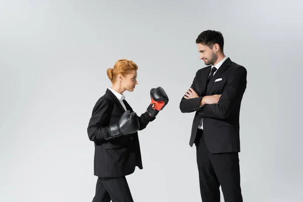 Улыбающийся бизнесмен, стоящий со скрещенными руками и смотрящий на рыжую деловую женщину в боксёрских перчатках, изолированных на сером — стоковое фото