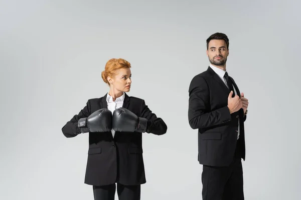 Redhead businesswoman in boxing gloves looking at haughty businessman in stylish blazer isolated on grey — Stock Photo