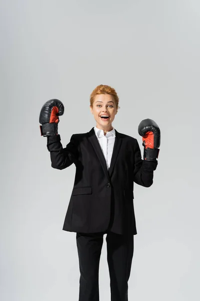 Aufgeregte rothaarige Geschäftsfrau in schwarzem Anzug und Latex-Handschuhen jubelt vereinzelt auf grau — Stockfoto