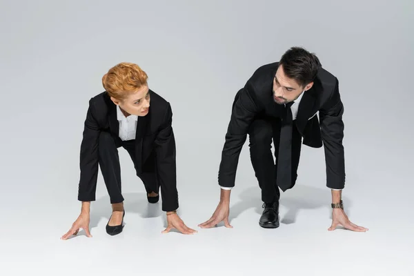 Full length of business competitors in black formal wear standing in low start position and looking at each other on grey background — Stock Photo