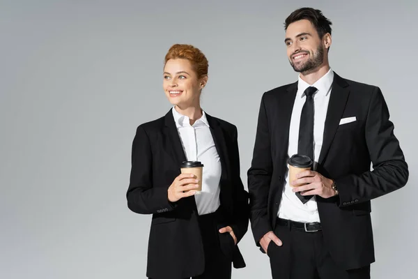 Alegres socios de negocios sosteniendo café para llevar de pie con las manos en los bolsillos y mirando hacia otro lado aislado en gris - foto de stock