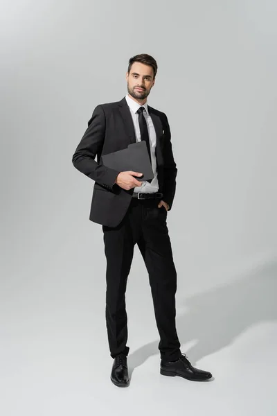 Повна довжина успішного і впевненого бізнесмена в чорному стильному костюмі, що стоїть з текою на сірому фоні — стокове фото