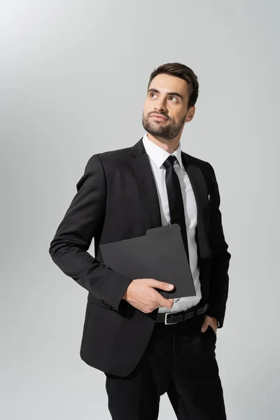 Вдумливий бізнесмен в чорному костюмі тримає папку і дивиться ізольовано на сірому — стокове фото