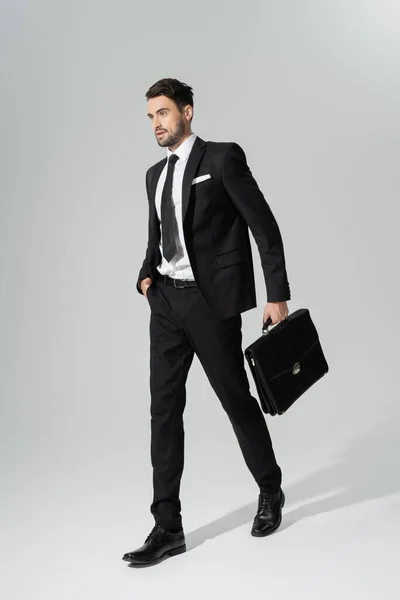 Full length of businessman in black pantsuit walking with briefcase on grey background — Stock Photo