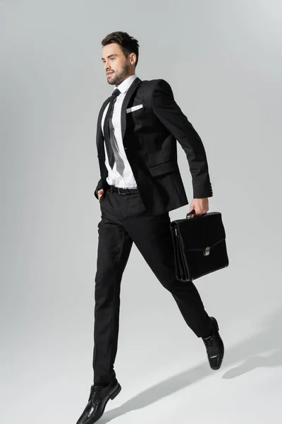 Longitud completa de joven hombre de negocios en traje pantalón negro corriendo con maletín y mano en bolsillo sobre fondo gris — Stock Photo