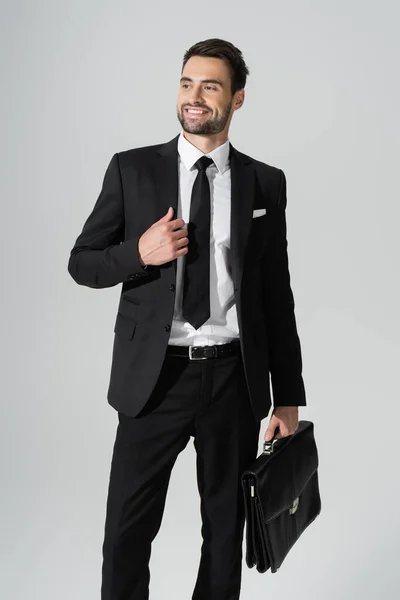 Радісний бізнесмен в чорному стильному костюмі, що стоїть з портфелем і дивиться ізольовано на сірий — стокове фото