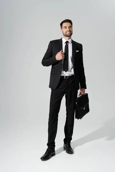 In voller Länge glücklicher erfolgreicher Geschäftsmann mit lederner Aktentasche, der auf grauem Hintergrund wegschaut — Stockfoto