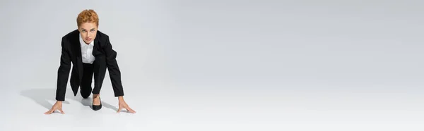 Voller Länge rothaarige Geschäftsfrau im schwarzen Strumpfanzug, die von niedriger Startposition auf grauem Hintergrund in die Kamera blickt, Banner — Stockfoto