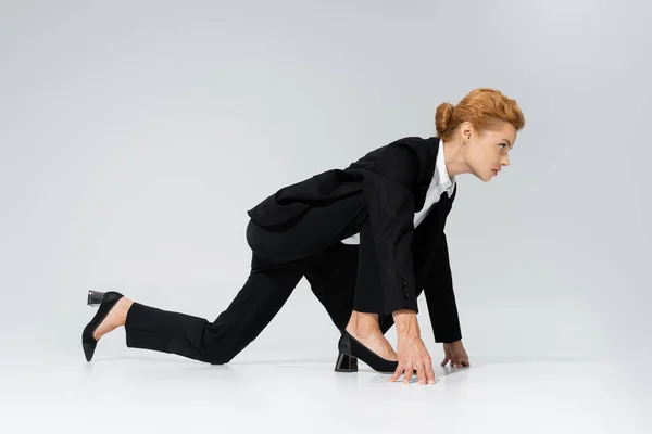 Вид збоку визначеної бізнес-леді в чорному костюмі, що стоїть в низькому стартовому положенні на сірому фоні — стокове фото