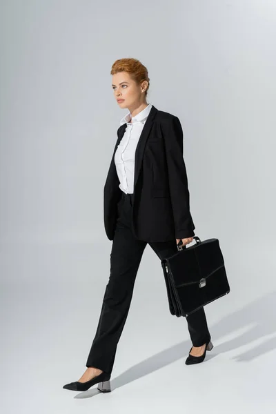 Pleine longueur de femme d'affaires confiante regardant vers l'avenir tout en marchant avec mallette sur fond gris — Photo de stock